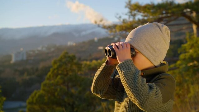 Boy looking through binoculars around while in nature, in the mountains in the rays of the setting sun on a spring evening.