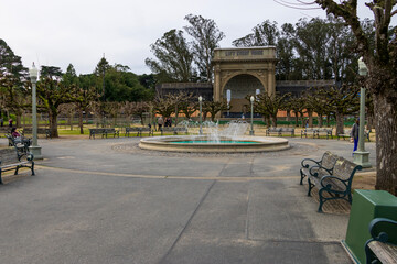 a circular water fountain in the center of the courtyard at Golden Gate Park with people walk and green trees, plants and grass in San Francisco California USA
