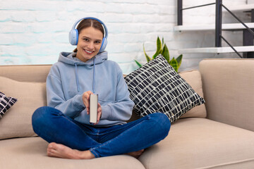 Woman listening music in headphones and reading book while sitting on couch.