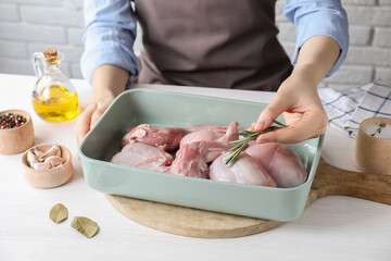 Woman putting rosemary into baking dish with raw rabbit meat at white wooden table, closeup