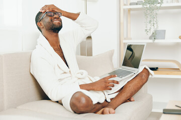 Smiling African American Freelancer Working on Laptop in a Modern Home Office, Typing and Enjoying the Morning The young black guy, dressed in a bathrobe, is sitting on a comfortable sofa in the