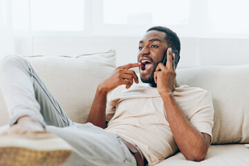Young African American man sitting on a black sofa at home, happily talking on his smartphone during a video call With a relaxed posture and casual tshirt, he confidently holds the phone and engages