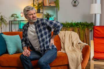 Tired senior bearded man suffering from back pain sitting on couch in living room at home. Sad old...