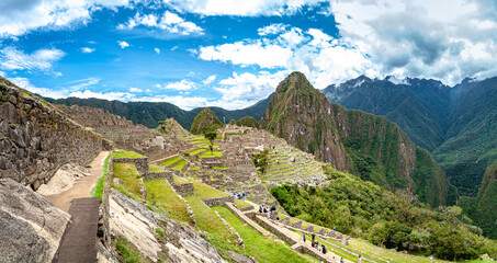 A complete scene of Macchu Pichu, in a blue sky day. Many tourists are present and scattered around the place visiting the ruins.