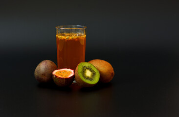 A tall glass of fruit juice with seeds on a black background, next to pieces of ripe kiwi and chopped passion fruit.