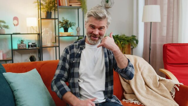 Senior Caucasian bearded old man pointing at camera doing phone gesture saying hey you call me back at home. Mature gray-haired old guy in living room sitting on sofa. Call me, here is contact number