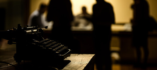 Silhouette of a vintage typewriter with blurred people in the background, depicting creative writing, journalism, or historical technology concept in a moody atmosphere, background with a place for te