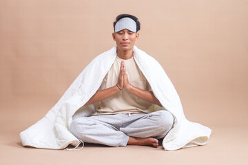 Young Asian man with eye mask and pajamas jam, sitting on floor and covered with blanket doing...