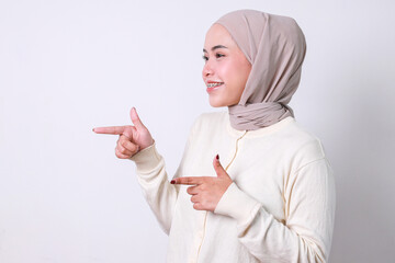 Asian muslim woman with braces pointing finger aside makes gun gesture, greets friend or approves idea, isolated over white background