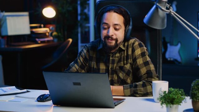 Cheerful man relaxing at home by watching TV show on laptop using headphones. Happy person enjoying leisure time in neon lit apartment at night, consuming entertainment media content, camera A