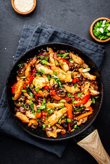 Hot stir fry chicken  slices with red paprika, mushrooms, chives and sesame seeds in frying pan. Black table background, top view