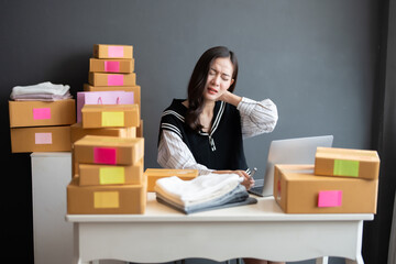 Asian female online seller is a business owner. Feeling tired and sore from working to order products for customers in the office. On the table were boxes ready for delivery and a laptop.