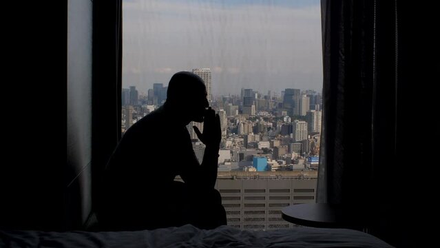 Silhouette Of Lonely Male Anxiously Sitting Near High Rise Apartment Window, Rocking Back Showing Loneliness, Thinking And Anxiety. Static Shot