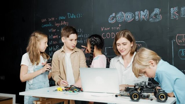 Caucasian teacher using laptop and explain about engineering system while diverse student holding electronic equipment in front of blackboard with prompt or code written in STEM class. Erudition.