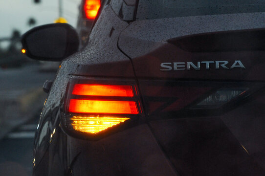 A close-up partial view of the rear end of a Nissan Sentra, showcasing the red brake light and amber/yellow left signal light in clear detail during rainy weather