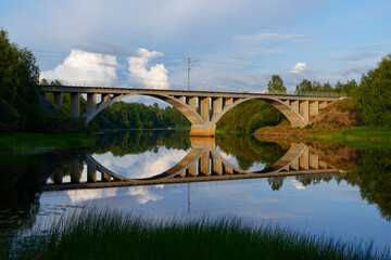 A railway bridge over calm water lit by the sun on a late summer evening with reflection on the water surface