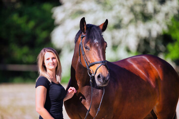 Young brunette woman in the sunshine with her horse in front of white flowering bushes.