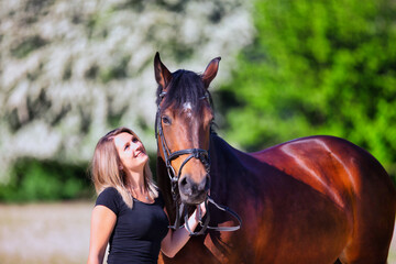 Obraz premium Young brunette woman in the sunshine with her horse in front of white flowering bushes.