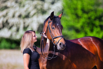 Obraz premium Young brunette woman in the sunshine with her horse in front of white flowering bushes.