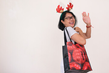 Asian woman wearing Christmas hair band, waving her hand with big shopping bag in her right shoulder