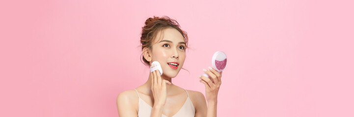 Beautiful young woman applying face powder with puff applicator on pink background