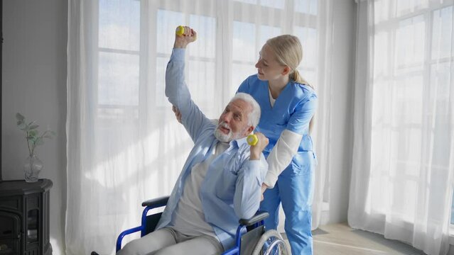 Man in wheelchair exercising with nurse. Social medical worker woman helping senior male doing physical hands exercises with dumbbell. Palliative care pensioner with neurodegenerative pathologies.