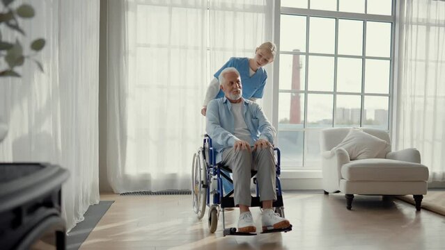 Social worker helps man in wheelchair. Female social welfare physical therapist looks after senior male, talking, providing medical care at hospital. Living with legs paralysis, special needs people.