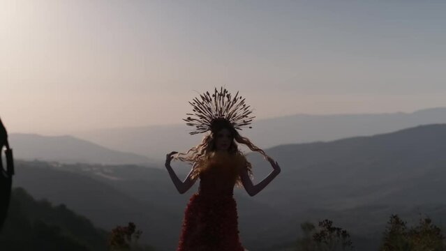 A young woman in an autumn outfit of flowers and grass and a headdress made of ears of corn stands against the background of the sunset in the mountains