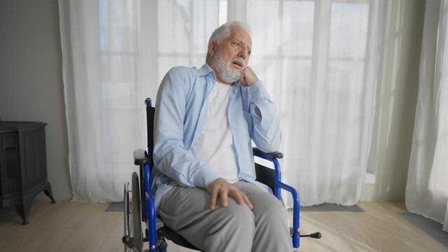 Old man in wheelchair looks at window. Thoughtful gray haired senior male at home enjoying pastime leisure. Living with physical legs trauma paralysis, sadness, loneliness of older people concept.