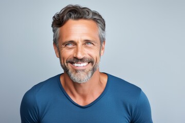 Handsome mature man in blue t-shirt smiling at camera while standing against grey background