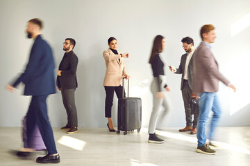 Obraz premium Young woman with a traveling suitcase standing by the wall and looking at her watch while waiting at the train station or airport among a blurred crowd of people walking by fast