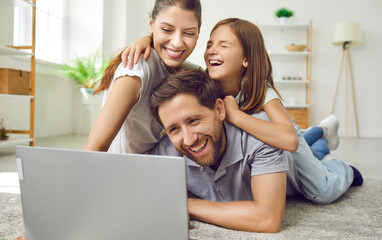 Portrait of happy loving family using laptop while spending time together at home in living room. Mom, dad and their daughter are lying on carpet and watching video, movie or browsing Internet.