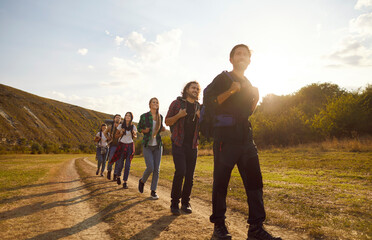 Young happy people tourists with large backpacks hiking in nature walking in a row in the mountains. Smiling friends having trekking tour outdoors. Adventure, travel and tourism concept.