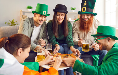 Young happy friends group sitting at home eating pizza and drinking beer at Saint Patrick's Day...