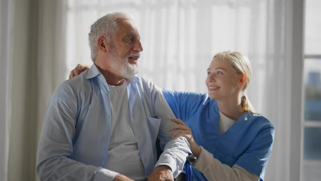 Nurse talking senior man in wheelchair. Female social welfare physical therapist looks after senior male, supporting, providing medical care at home. Living with legs paralysis, special needs people.