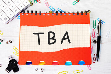TBA (abbreviation of to be announced) word is written on a sheet in a cage lying on a notebook on the table next to stationery - obrazy, fototapety, plakaty