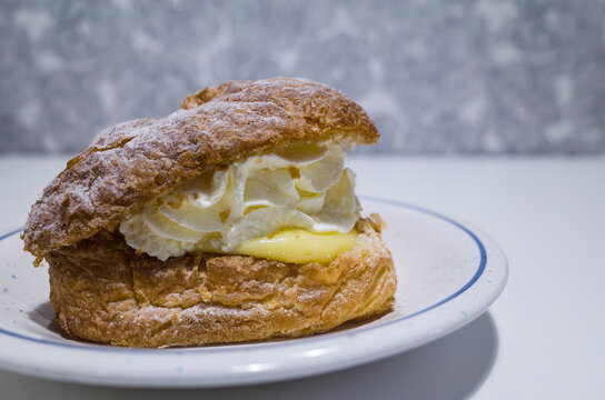 Swedish wiener semla, shrove bun made of puff pastry filled with whipped cream and vanilla cream, close up of sweet dessert on a plate on white table by the gray wall
