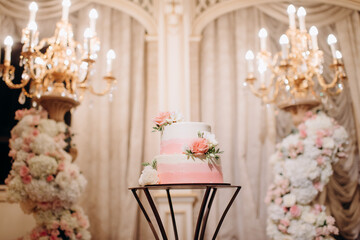elegant white two-tiered wedding cake decorated with flowers on a small table against the backdrop of a wedding arch
