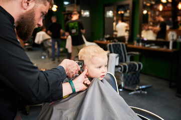 Barber using comb and shaver to cut hair. Professional hairdresser shaving little kid's nape.