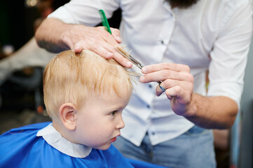 Man barber cutting little boy's hair using comb and scissors. Child getting haircut from adult...
