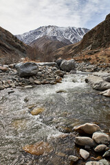 Early spring natural landscape. Snow capped mount, hillsides, large boulders and rocky bank of a shallow river with fast flowing water. Kyrgyz Republic Kyrgyzstan mountains