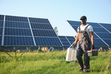 African american man in white helmet and grey overalls standing among rows of solar panels