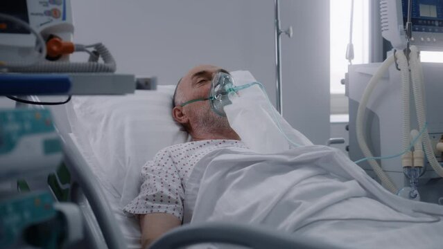 Senior man in oxygen mask lies in bed in hospital ward. Old patient during artificial lung ventilation. Modern emergency room in clinic. Intensive care coronavirus department in medical facility.