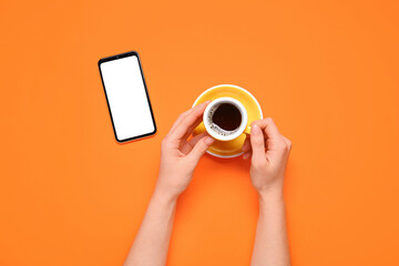 Female hands holding cup of coffee and smartphone with blank screen on orange background