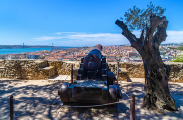 Restored bronze cannon from St. George's Castle overlooking the city of Lisbon and the red metal 25 de Abril suspension bridge next to an olive tree.