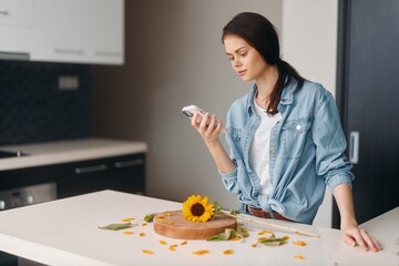 Beautiful young woman sitting at the kitchen table, holding a mobile phone and enjoying a healthy breakfast