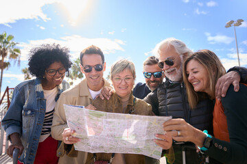Group of diverse middle-aged tourist people standing looking at travel map in hands on street of vacation city happy and smiling on sunny day. Adults friends in community enjoying a trip together  - obrazy, fototapety, plakaty