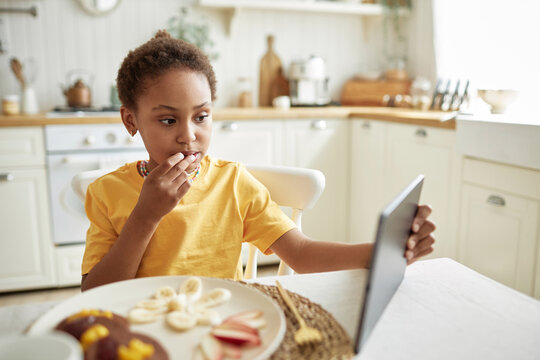 Selective focus shot of modern African American girl sitting at table in kitchen eating breakfast and watching videos in Internet on digital tablet