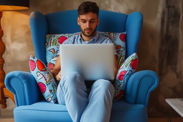 man sitting in a blue armchair with pop art cushions, laptop on lap