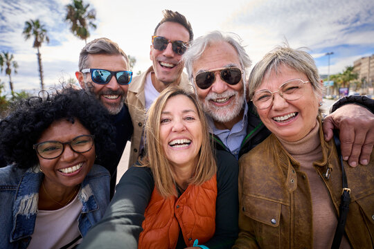 Group of diverse cheerful middle-aged tourist friends posing smiling taking a photo selfie embracing together with front camera on travel outdoor. Mature happy six people enjoy holidays sunny day 